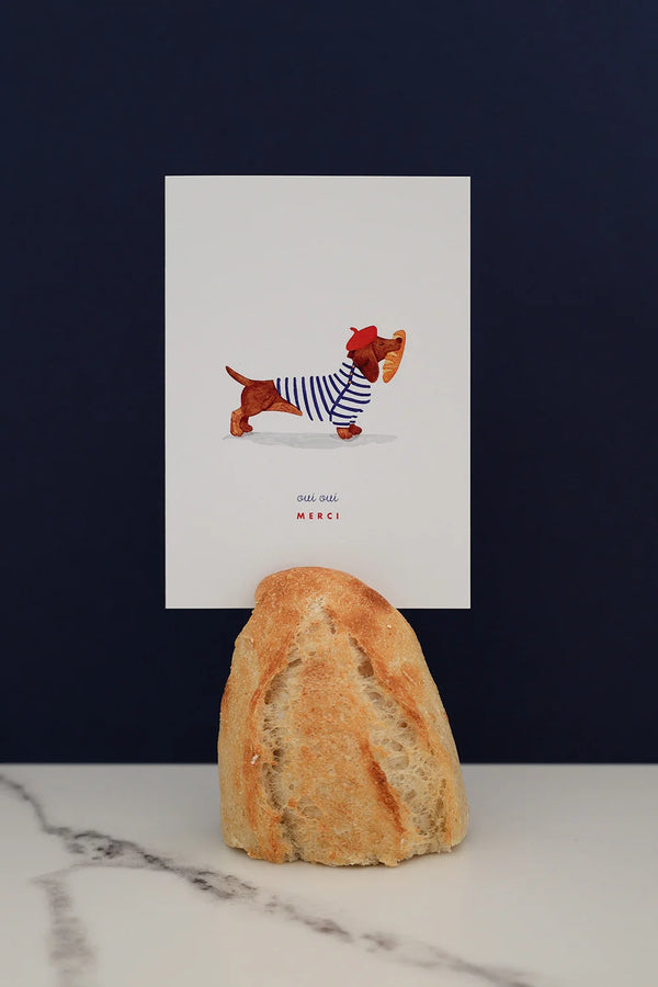 All The Ways To Say Oui Oui Merci card – dachshund in striped shirt holding baguette, styled with bread and marble backdrop