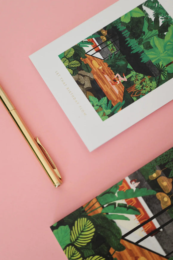 Birthday card featuring a tropical yoga scene with a woman practicing, styled on a pink background with a gold pen.