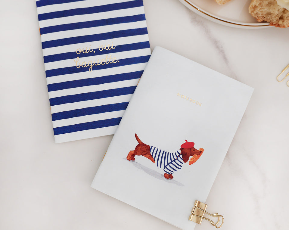 Set of two premium notebooks featuring a whimsical dachshund in a beret carrying a baguette and a classic blue striped design with gold foil accents.