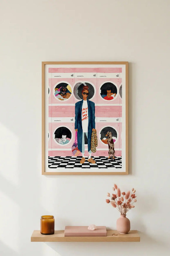 Minimalist interior mockup featuring the "Laundry Day" art print in a natural light wood frame hanging on a plain white wall. The stylish hand-painted illustration portrays a woman in a long blue coat, sunglasses, and a "Laundry Day Outfit" t-shirt standing inside a pink laundromat with a checkered floor, where cats and dogs are cozying up inside the washing machines. Below the poster, a floating wooden shelf holds an amber candle glass, a pink book, and a pink ceramic vase with fluffy dried flowers.