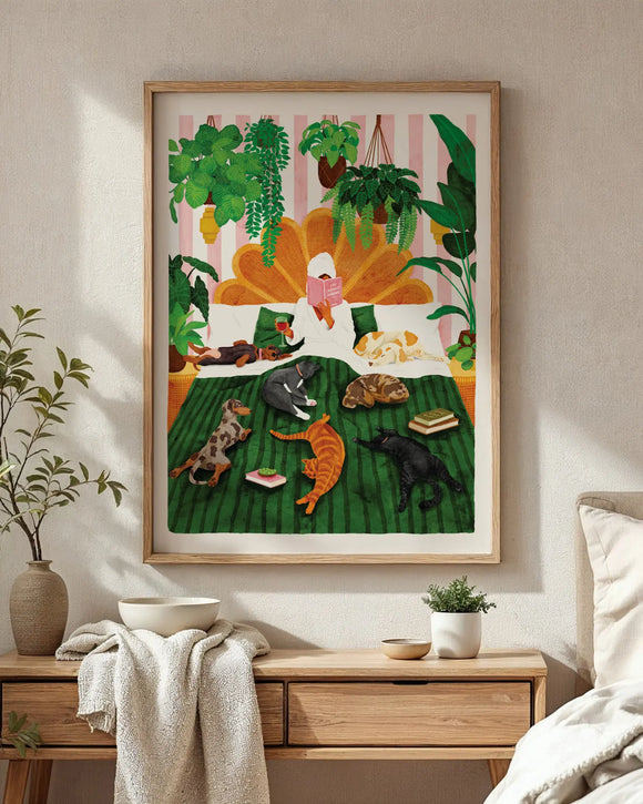 A bright bedroom interior mockup featuring the "Life with plants & pets" art print in a natural light wood frame hanging on a beige textured wall. The hand-painted illustration portrays a relaxing scene of a woman in a white bathrobe reading a book in bed, surrounded by lush hanging indoor plants and seven sleeping dogs and cats on a green striped blanket. Below the poster sits a light wooden dresser decorated with a ceramic bowl, a soft beige throw blanket, and a small potted green plant under warm sunligh