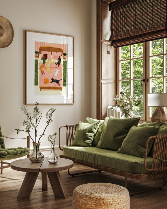 A cozy living room interior mockup featuring the "Happy pets" poster in a light wood frame hanging on a beige wall. The illustration shows a woman and her five happy pets—dogs and cats—relaxing together on a large pink bed. The room is decorated in a warm, natural style with a green velvet sofa, a rustic wooden coffee table with white flowers in glass vases, a woven jute pouf, and a large window with bamboo blinds overlooking lush green trees.