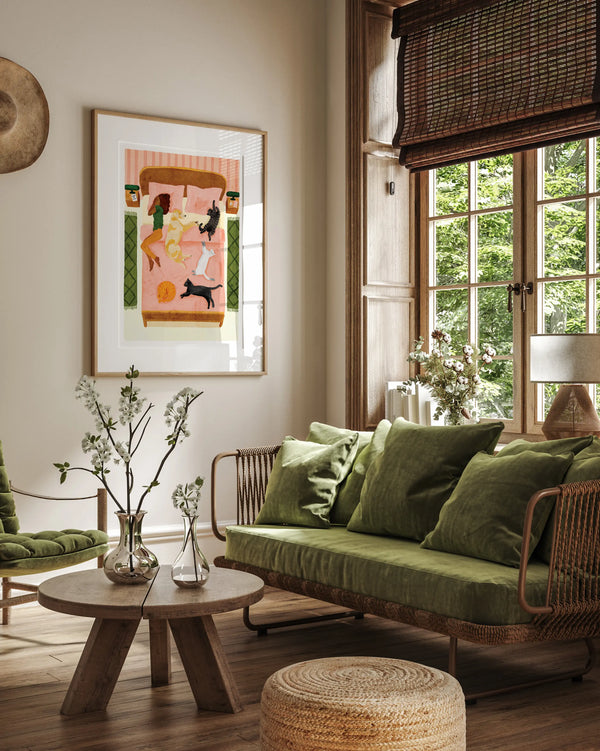 A cozy living room interior mockup featuring the "Happy pets" poster in a light wood frame hanging on a beige wall. The illustration shows a woman and her five happy pets—dogs and cats—relaxing together on a large pink bed. The room is decorated in a warm, natural style with a green velvet sofa, a rustic wooden coffee table with white flowers in glass vases, a woven jute pouf, and a large window with bamboo blinds overlooking lush green trees.