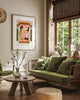 A cozy living room interior mockup featuring the "Happy pets" poster in a light wood frame hanging on a beige wall. The illustration shows a woman and her five happy pets—dogs and cats—relaxing together on a large pink bed. The room is decorated in a warm, natural style with a green velvet sofa, a rustic wooden coffee table with white flowers in glass vases, a woven jute pouf, and a large window with bamboo blinds overlooking lush green trees.