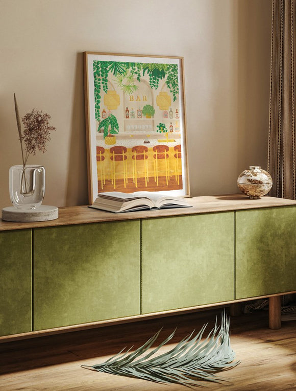 Light wood framed Bar art print resting on a modern olive green sideboard with a glass vase and an open book.
