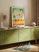 Light wood framed Bar art print resting on a modern olive green sideboard with a glass vase and an open book.