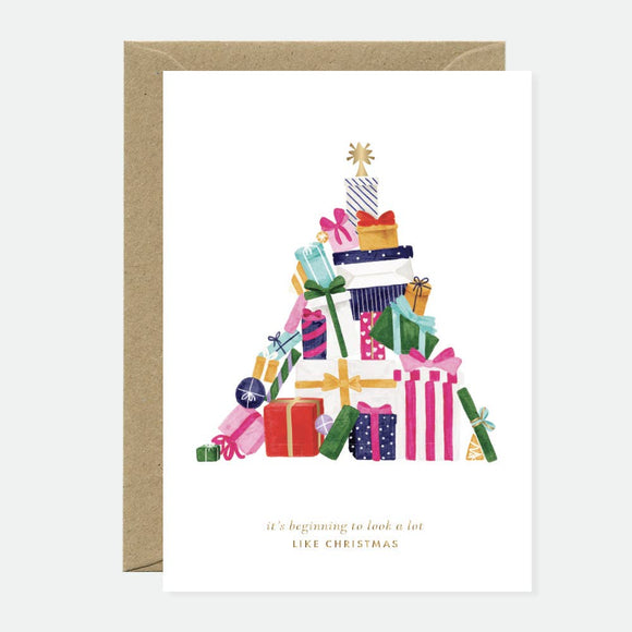 A greeting card with a gold foil design featuring a Christmas tree shape, filled with various presents, and a blank interior. The card is accompanied by a recycled brown envelope.
