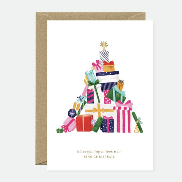 A greeting card with a gold foil design featuring a Christmas tree shape, filled with various presents, and a blank interior. The card is accompanied by a recycled brown envelope.
