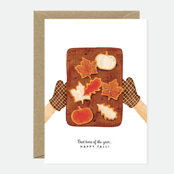 Greeting card showing autumn cookies shaped like pumpkins and colorful fall leaves on a wooden tray with oven mitts, evoking cozy seasonal vibes.