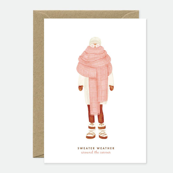 Greeting card with an illustration of a person wearing a pink scarf and hat, and text 'Sweater Weather' alongside. The card has a blank interior and comes with a brown envelope.