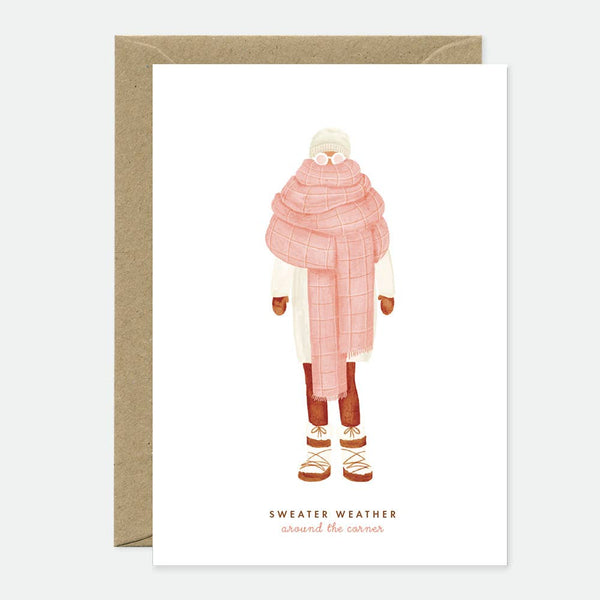 Greeting card with an illustration of a person wearing a pink scarf and hat, and text 'Sweater Weather' alongside. The card has a blank interior and comes with a brown envelope.