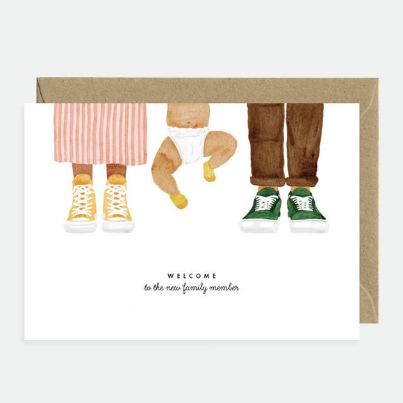 Greeting card with an illustration of a baby wearing sneakers and a text that reads 'Welcome Baby Family Member'. The card has a natural white background and a recycled brown envelope.