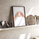 Framed Hands of love poster leaning on a floating ledge alongside artistic paintbrushes and a round woven tray.