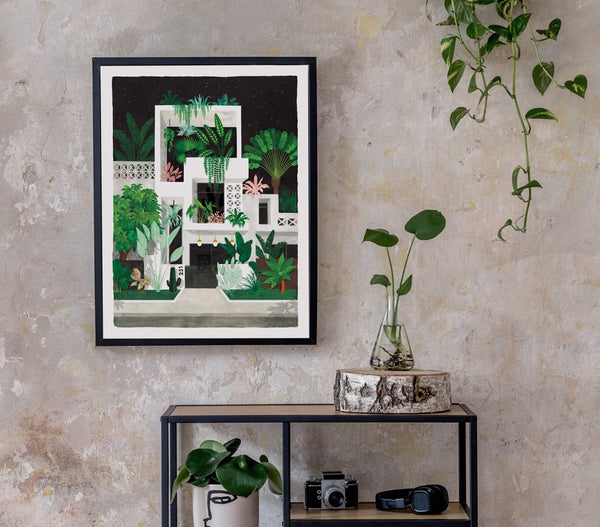 Framed Miami by night art print hanging on a rustic concrete wall above a metal shelf with indoor plants and a vintage camera.