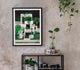 Framed Miami by night art print hanging on a rustic concrete wall above a metal shelf with indoor plants and a vintage camera.