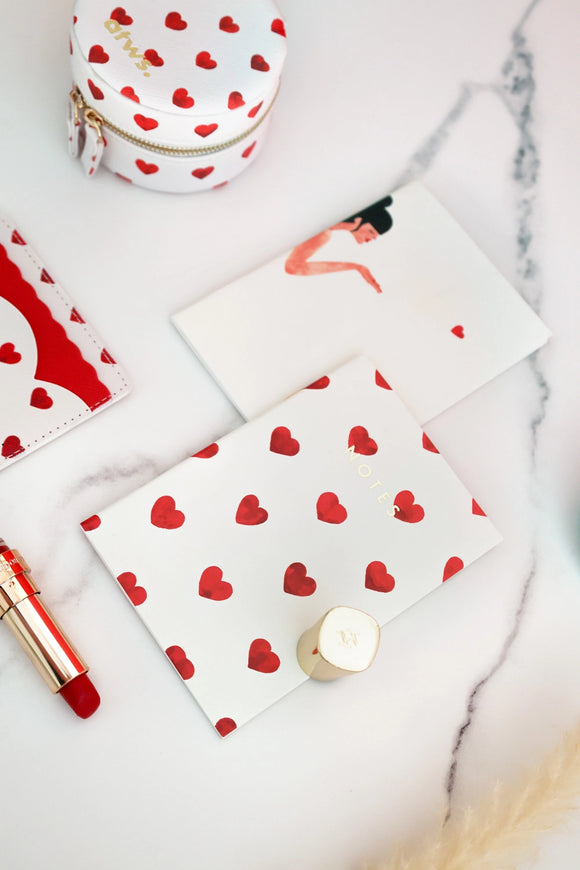 Notebook with red heart pattern on white cover styled with lipstick, pouch, and accessories on marble surface.