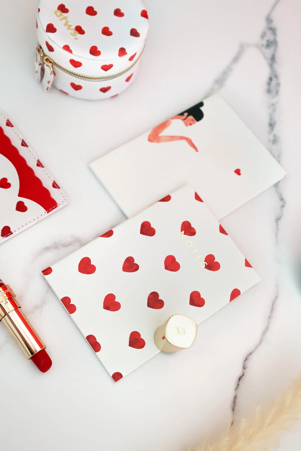 Notebook with red heart pattern on white cover styled with lipstick, pouch, and accessories on marble surface.