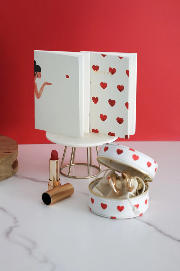 Romantic notebook with red heart design on white cover, displayed with jewelry case and lipstick against red background.