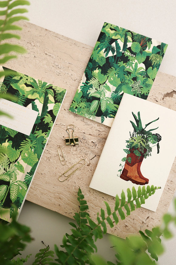 Botanical notebooks with green leafy designs and boot illustration, styled with paper clips and surrounded by fresh plants.