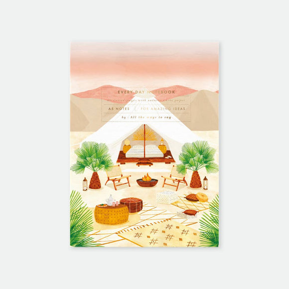 A5 notebook with a gold foil cover featuring an illustration of a desert scene with a tent, palm trees, and campsite furniture.