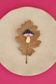 Crazy Mushroom enamel pin by All The Ways To Say on brown oak leaf over beige plate – pink background