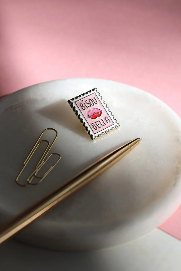 Bisou Bella enamel pin photographed on white marble tray with gold pen – artistic lighting and soft pink background