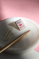 Bisou Bella enamel pin photographed on white marble tray with gold pen – artistic lighting and soft pink background