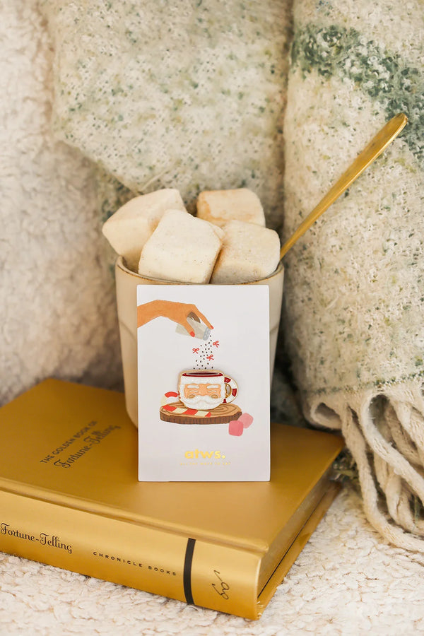 Santa’s Cup enamel pin by All The Ways To Say on illustrated card inside cozy winter setup with marshmallows and blanket