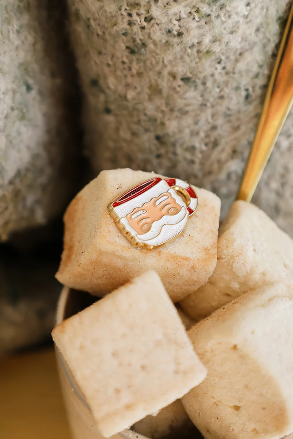 Santa’s Cup enamel pin by All The Ways To Say held in hands around a mug of marshmallows – soft and comforting winter vibe