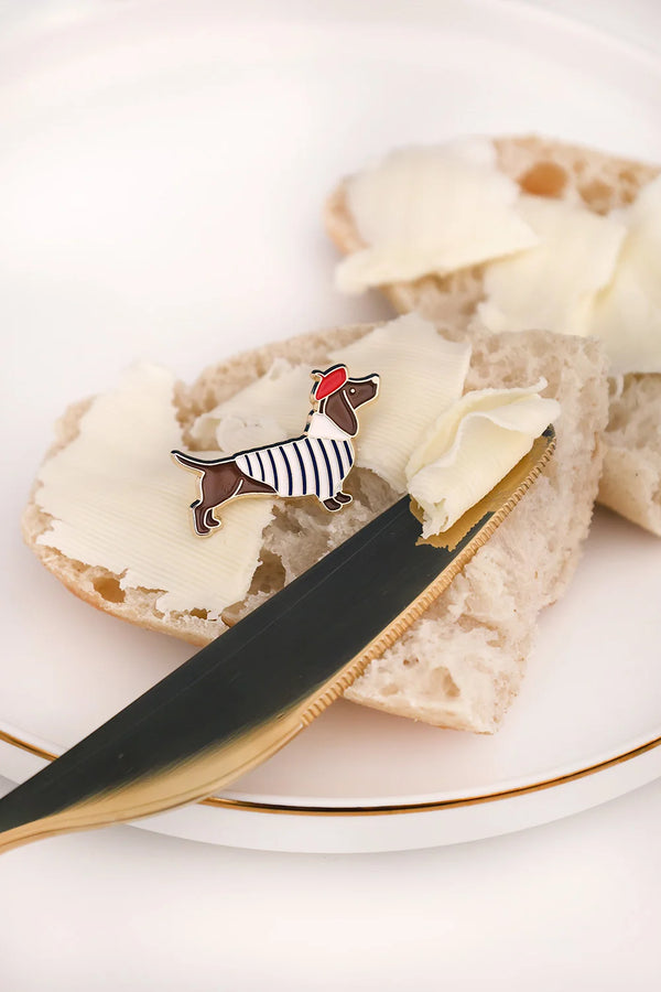 Little Frenchie enamel pin by All The Ways To Say on slice of bread with butter and gold knife – French lifestyle flatlay