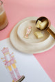 Pink Nutcracker enamel pin by All The Ways To Say on beige ceramic plate with white chocolate pieces – festive pastel setup
