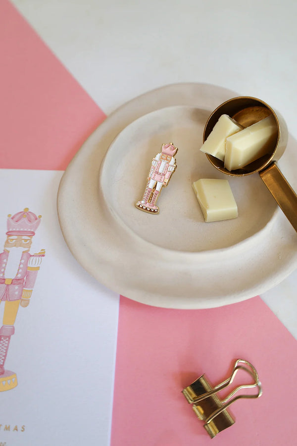Pink Nutcracker enamel pin by All The Ways To Say on pink and white background – cozy Christmas mood with chocolate and gold details