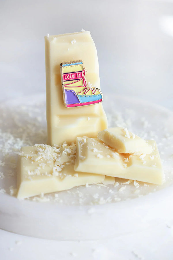 Close-up of Cold AF enamel pin by All The Ways To Say – pastel snow boot on creamy chocolate bar surface