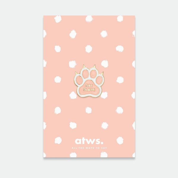 A gold plated enamel pin in the shape of a cat paw, displayed on a card with a pink polka dot background.