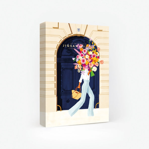 1000-piece jigsaw puzzle box by All The Ways To Say featuring a woman carrying a massive floral bouquet in front of a grand navy blue Parisian door.