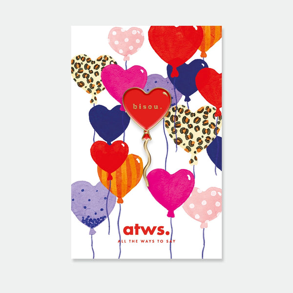 A red heart balloon enamel pin by All The Ways To Say with "bisou." engraved in gold, displayed on a colorful heart-themed card.