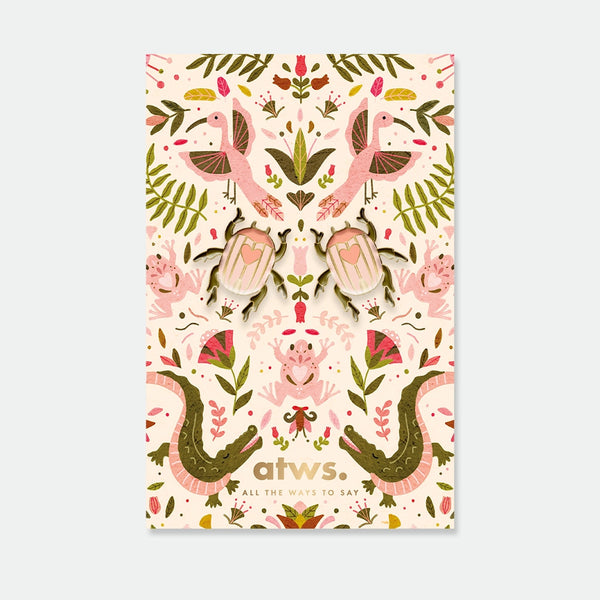 A set of two symmetrical pink beetle enamel pins with gold hearts on their backs, displayed on a lush botanical backing card.