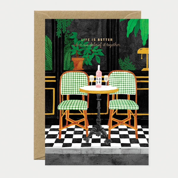 A sophisticated greeting card by All The Ways To Say featuring a Parisian bistro scene with two green checkered chairs, a bottle of rose wine, and the text "LIFE IS BETTER when we debrief it together."