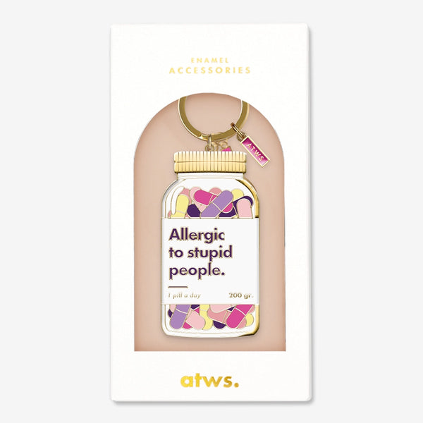 Un porte-clés humoristique en émail doré All The Ways To Say en forme de flacon de pilules avec le texte "Allergic to stupid people.", présenté dans son étui.