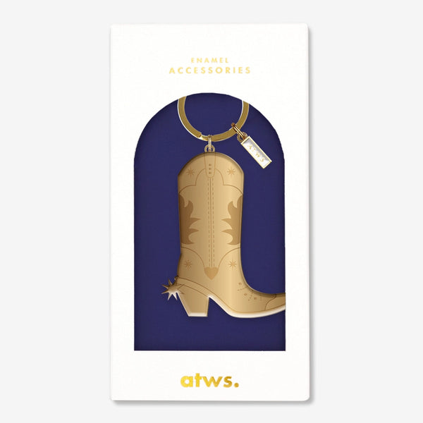 A stylish gold enamel keychain by All The Ways To Say in the shape of a cowboy boot, presented in an elegant navy blue packaging.