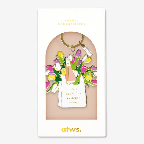 A stylish gold enamel keychain by All The Ways To Say featuring a white bag with a rosé bottle and tulips, inscribed with "it's a great day to drink rosay."