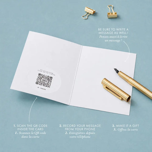 Inside view of an audio greeting card open flat, showing a QR code sticker on the left page and a blank right page for a handwritten message, with a gold pen and binder clips on a blue-grey background