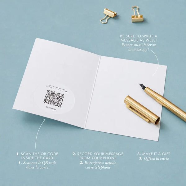 Inside view of an audio greeting card open flat, showing a QR code sticker on the left page and a blank right page for a handwritten message, with a gold pen and binder clips on a blue-grey background