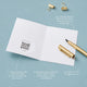 Inside view of an audio greeting card open flat, showing a QR code sticker on the left page and a blank right page for a handwritten message, with a gold pen and binder clips on a blue-grey background