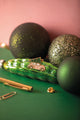 Christmas car enamel pin, a pink vintage car carrying a Christmas tree on its roof, among green glitter baubles