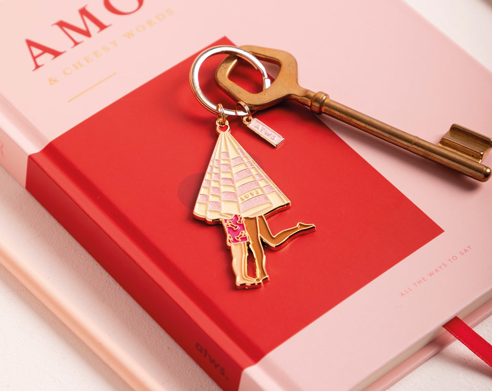 Charming enamel keychain featuring a romantic couple under a pink umbrella, resting on a red hardcover notebook next to a vintage brass key.