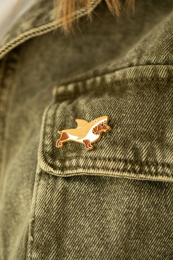 Shark Attack enamel pin, a dachshund in a shark costume with heart sunglasses, pinned on an olive denim jacket pocket
