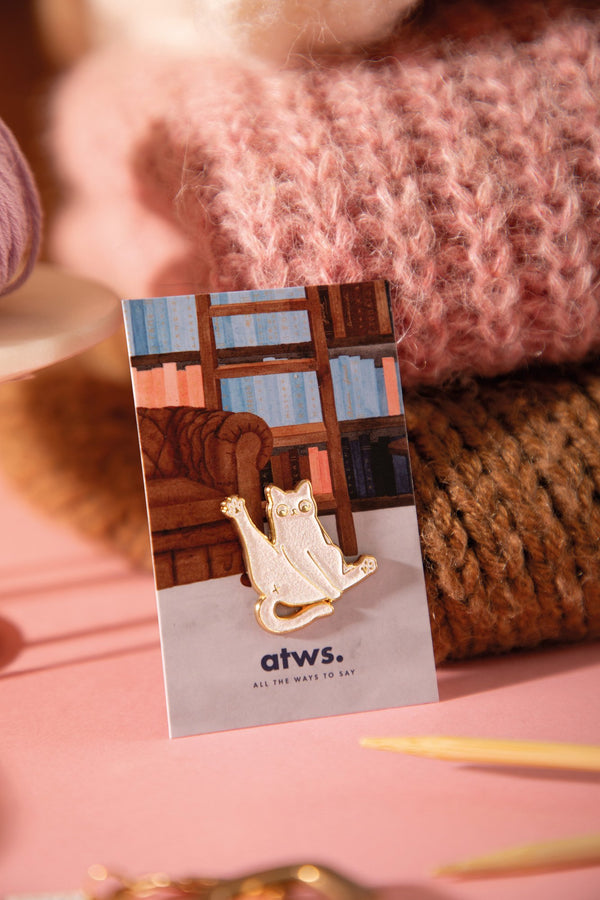 "Don't give a sh*t" white cat enamel pin with one paw raised, on an illustrated library backing card, against knitted wool