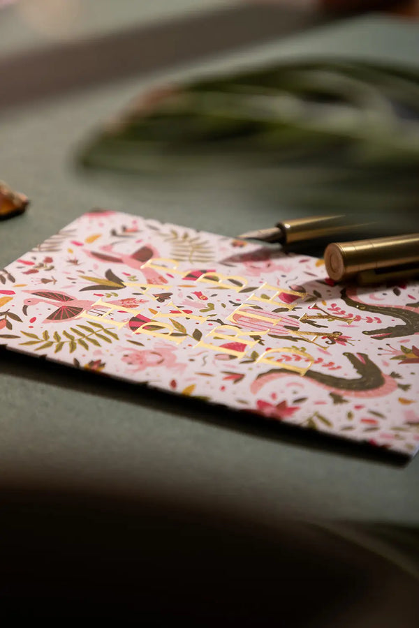 Happy birthday pink croco Greeting card - Close-up view on a dark green desk with gold stationery accessories.