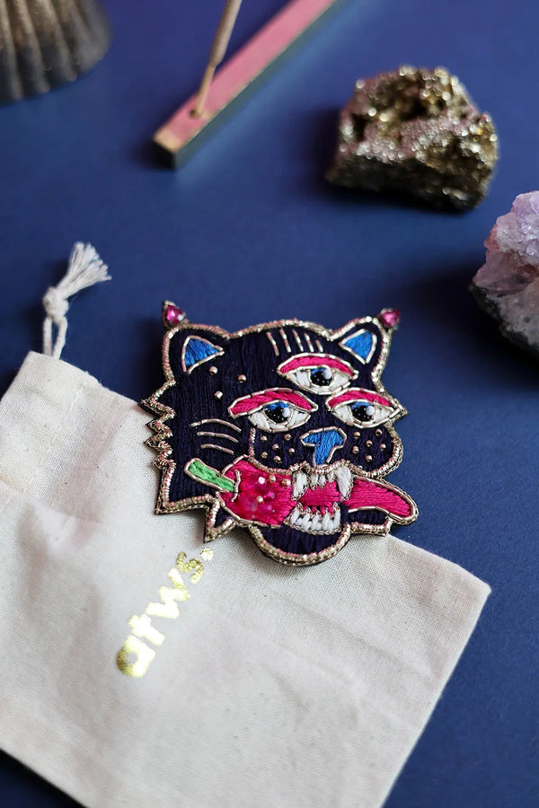 Psychotropic tiger embroidery pin displayed next to a cream cotton jewelry pouch with gold branding on a blue surface.